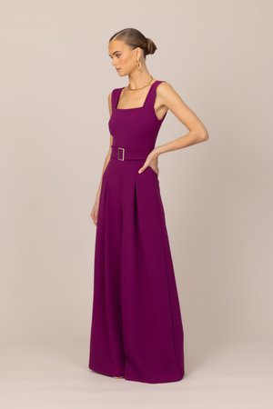 Moteris vilki be rankovių, purpurinės spalvos kombinezoną su diržu ir plačiomis kelnėmis, stovinti šonu, plaukus sukusi į žemą kuodą, dėvi auksinius auskarus.