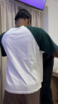 Ένα άτομο φοράει ένα πράσινο και άσπρο κοντομάνικο πουκάμισο ενώ κρατάει μαύρα παντελόνια μπροστά από μπεζ κουρτίνες και μια ανοιχτόχρωμη ξύλινη επιφάνεια.