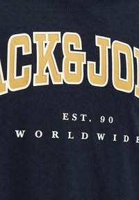 Tamsiai mėlynas medvilninis marškinėliai su dideliu geltonu tekstu "BLACK&JOHN" ir mažesniu baltu tekstu "EST. 90 PAAUGUSIAI KAS TIK." Tekstas turi drąsų, sluoksniuotą dizainą.