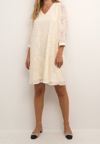 Hvid blondekjole med V-udskæring, trekvart lange gennemsigtige ærmer og en afslappet pasform. Udsmykket med blomster mønstre og en lige kant.