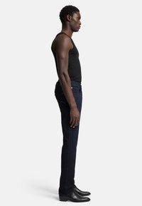 Jeans in denim scuro con una vestibilità slim, caratterizzati da una sottile accentuazione blu nei punti e un design di tasche standard, abbinati a una canotta nera.