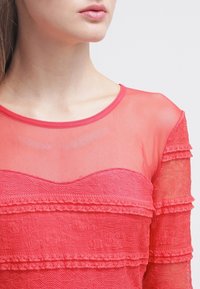 Top rojo transparente con rayas de encaje horizontales, escote redondo y tejido texturizado. Diseño suave y en capas con detalles delicados.