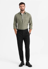Camicia button-up verde chiaro sopra una maglietta a righe bianca e nera, abbinata a pantaloni neri plissettati e scarpe nere stringate.