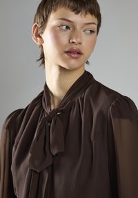 Braune, leichte Bluse mit langen, transparenten Ärmeln und einem gebundenen Ausschnitt. Subtile Textur und geraffte Details an den Schultern werten das Design auf.