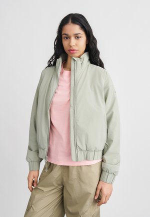 Ung kvinde med langt sort hår iført en lysegrå lynlåsjakke over en pink trøje og beige cargobukser, stående mod en ensfarvet baggrund.