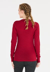 Kvinna med flätat hår som bär en figurnära långärmad röd tröja och mörkgrå byxor, stående med ryggen mot kameran mot en vit bakgrund.