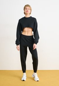 Musthave musta lühikeste varrukatega kapuutsiga pusake, mis on kombineeritud mustade kitsaste dressipükstega. Mudel kannab valgeid tossusid hallide aktsentidega.