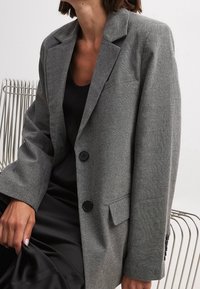 Femme assise sur une chaise en métal, portant un blazer gris ample sur un haut noir et une jupe noire, la tête partiellement hors du cadre.