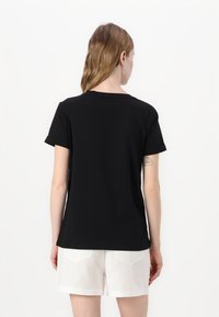 T-shirt nera a maniche corte, design a collo rotondo, texture liscia, indossata da un modello con pantaloni corti bianchi, sfondo neutro.