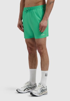 Mann trägt grüne Shorts mit Kordelzug, weiße Sportsocken mit schwarzem Schriftzug und silberne Laufschuhe, steht vor einem schlichten Hintergrund.
