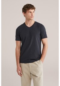 Jaunuolis dėvi juodą V formos kaklo marškinėlį ir smėlio spalvos kelnes, stovi viena ranka kišenėje, žiūrėdamas į šoną.