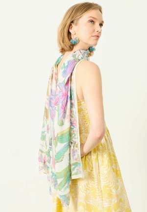 Frau mit kurzem blonden Haar, die blaue blumenförmige Ohrringe, ein gelbes Kleid mit Blumenmuster und einen pastellfarbenen Blumenschal über der Schulter trägt.