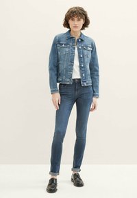 Giacca di jeans blu medio con tasche frontali e bottoni argentati, indossata sopra una maglietta bianca e jeans skinny blu scuro. Scarpe nere con lacci.