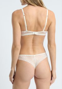 Ensemble de soutien-gorge et string en crème, avec des bords festonnés et des motifs floraux. Bretelles réglables et un détail en nœud au dos.