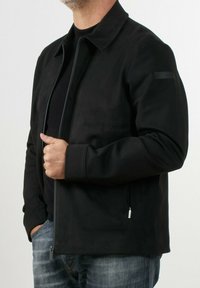 Giacca nera con colletto alto, tessuto morbido, chiusura con zip, tasche laterali e branding discreto sulla manica. Progettata con una vestibilità su misura.
