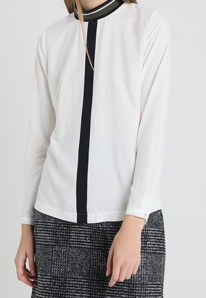 Femme portant un chemisier blanc à manches longues avec une bande noire verticale et un col noir, assorti à une jupe à carreaux grise.