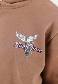 Brun hoodie med en grafisk design av en örn och blommor, med texten "Sixth June" i rosa, på ett mjukt, strukturerat tyg.