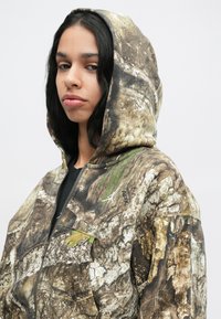 Ung person med langt sort hår iført en brun og grøn camouflage zip-up hoodie med hætten oppe, tydeligt drejet lidt til venstre mod en ensfarvet baggrund.