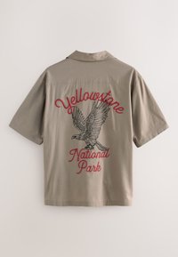 Chemise kaki à manches courtes avec un motif d'aigle au dos et un texte rouge brodé : « Yellowstone National Park ». Conception classique du col.