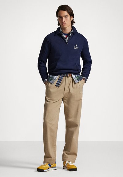 Pullover da marinha com fecho e logótipo, sobre uma camisa às riscas, combinado com calças bege e sapatilhas amarelas com acentos azuis.