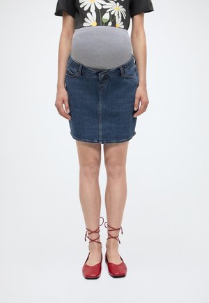 Tehotná osoba nosiaca sivý tehotenský pás pod čiernym tričkom s margarétkami, rifľovou sukňou a červenými topánkami na šnurovanie, stojaca na bielom pozadí.