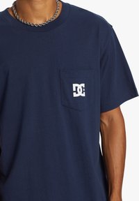 Námorně modré bavlněné tričko s krátkými rukávy, detailem kapsy a bílým logem na levé straně hrudi. Hladká textura a volný střih.