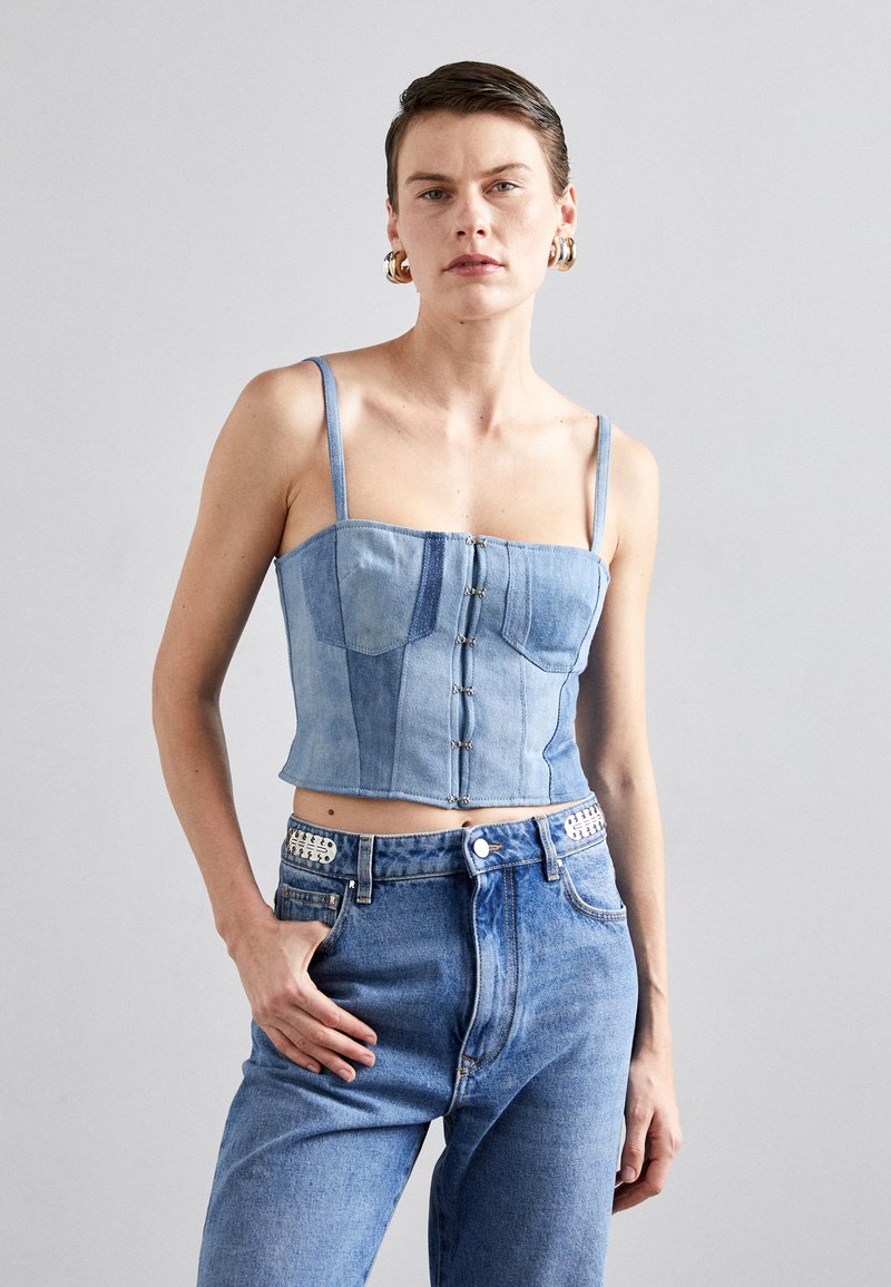 Lys blå denim crop top med en tettsittende design, tynne stropper, knapplukking foran og paneldetaljer. Båret med matchende høyt liv jeans.