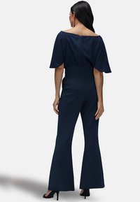 Marineblauwe jumpsuit met uitlopende benen en een brede halslijn. Heeft korte, vloeiende mouwen en een verborgen ritssluiting aan de achterkant. Gladde textuur van de stof.