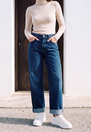 Une personne portant un col roulé crème, un jean bleu à revers et des baskets blanches se tient devant une porte en bois, les mains dans les poches.
