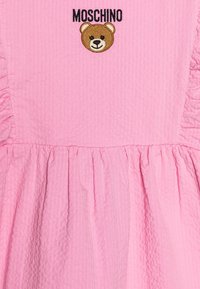Vestido rosa con textura fruncida, detalles con volantes y bordado del logo del oso Moschino en el pecho. Diseño casual, tejido ligero.
