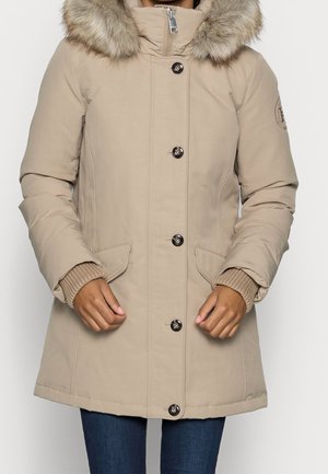 Kobieta w beżowej, pikowanej zimowej kurtce z kapturem obszytym futrem, zapinanej na guziki i zamek błyskawiczny, z klapowymi kieszeniami i ściągaczami na mankietach, widziana od połowy ud w górę.