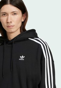 Čierna mikina s kapucňou s bielymi trojpruhmi na pleciach a malým bielym logom Adidas na hrudníku. Mäkká látka, kapucňa so sťahovacou šnúrkou.