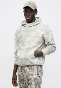 Felpa con cappuccio di un beige chiaro con motivo a foglie, dotata di tasca a marsupio. Abbinata a pantaloni coordinati con design e schema di colori simili.