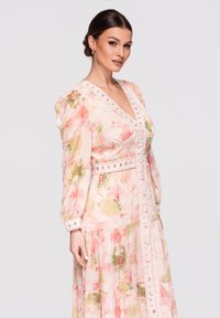Femme aux cheveux foncés coiffés en chignon bas, portant une robe pastel à manches longues à motif floral, avec un bord en dentelle et des boutons sur le devant.
