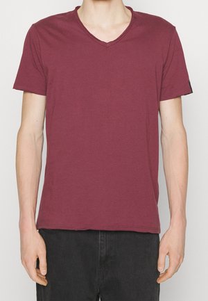 Homme portant un T-shirt uni bordeaux à col en V et manches courtes, ainsi qu'un pantalon noir, debout avec les bras détendus le long du corps, devant un fond neutre.