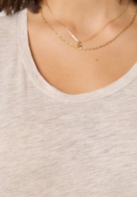 Ženska, ki nosi dve nežni zlatini ogrlici z verižico čez svetlo bež majico z okroglim izrezom.