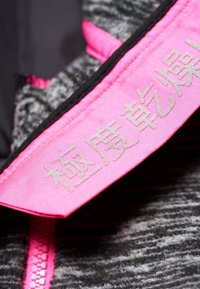 Veste en tissu gris texturé avec un col rose. Le col présente des lettres embossées en argent. Dispose d'une fermeture éclair rose.