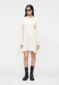 Smėlio spalvos ilgas marškinėlių tipo suknelė ilgomis rankovėmis ir sagomis priekyje, su siaura liemens dalimi ir platėjančia sijono dalimi. Dėvima su juodais karo stiliaus batais ir akiniais nuo saulės.