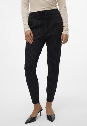 Vrouw draagt zwarte, op maat gemaakte broek met krijtstrepen, beige lange mouwen top en zwarte hooggehakte mules met spitse neus, staand tegen een witte achtergrond.