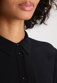 Svart skjorta med krage i ett strukturerat tyg, med tre synliga svarta knappar längs stängningen och en slät halsringning.