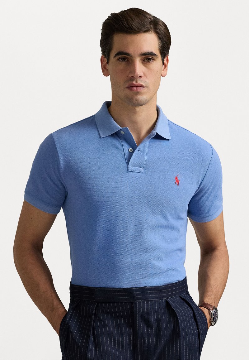Camisa polo azul claro en un tejido texturizado con un cuello clásico, que presenta un pequeño logo rojo en el lado izquierdo del pecho, combinada con pantalones de rayas.