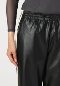 Sorte faux skinnbukser med en skinnende tekstur, elastisk midjebånd og kremfargede sidestiper. Nærbilde av buksene med en hånd plassert på stoffet.