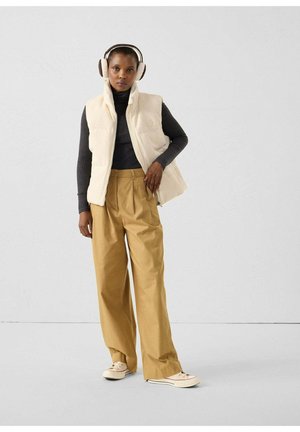 Chaleco crema sobre un suéter de cuello alto oscuro, combinado con pantalones sueltos de color marrón claro. El modelo lleva auriculares y zapatillas beige, y está de pie frente a un fondo claro.