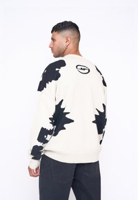 Pulover oversized de culoare crem, cu un model abstract negru și logo pe spate. Confecționat dintr-un material moale, cu manșete și tiv riglate.