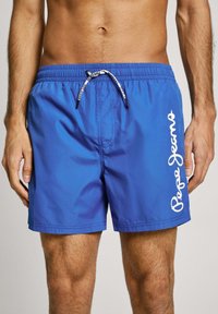 Μπλε μαγιό κατασκευασμένο από λείο συνθετικό υλικό, με ελαστική μέση, κορδόνι και λευκή εκτύπωση λογότυπου στο πλάι.