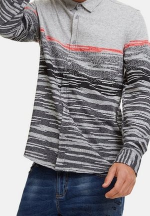 Uomo con camicia a maniche lunghe abbottonata con strisce astratte grigie, nere e rosse abbinata a jeans blu.