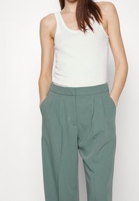 Débardeur blanc ajusté associé à un pantalon taille haute plissé de couleur vert pâle, avec poches latérales et texture lisse.