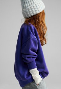 Fioletowa, oversize'owa bluza z prążkowanymi mankietami, zestawiona z białą, prążkowaną koszulą z długim rękawem. Model ma na sobie jasnoniebieską, dzianą czapkę beanie.