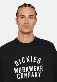 Black sweatshirt with a round neckline, featuring white text "DICKIES WORKWEAR COMPANY" in bold, curved letters across the front.