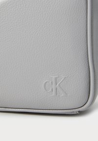 Grå lædertaske med tekstureret overflade, der har et præget "CK"-logo, rene linjer og stikninger for et raffineret udseende.
